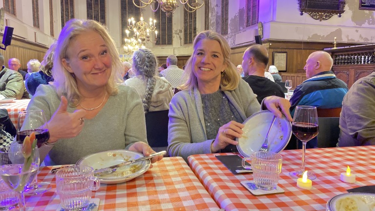 Twee lachende vrouwen aan een tafel met rood-wit geblokt tafelkleed, elk met een leeg bord en glazen wijn. Op de achtergrond is een ouderwetse zaal te zien.