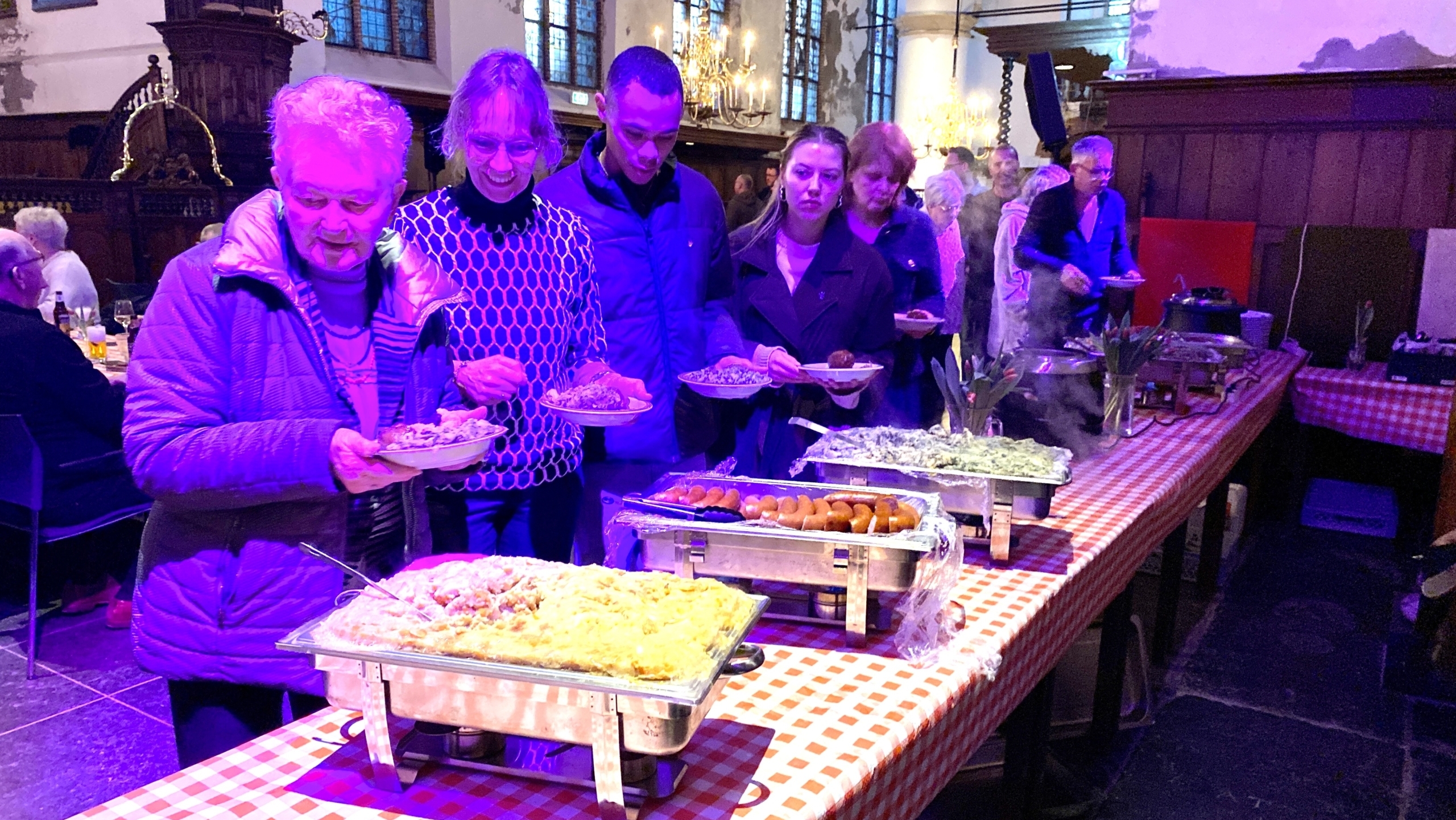 Mensen in de rij bij een buffet in een kerk met gedekte tafels en voedsel op schalen.