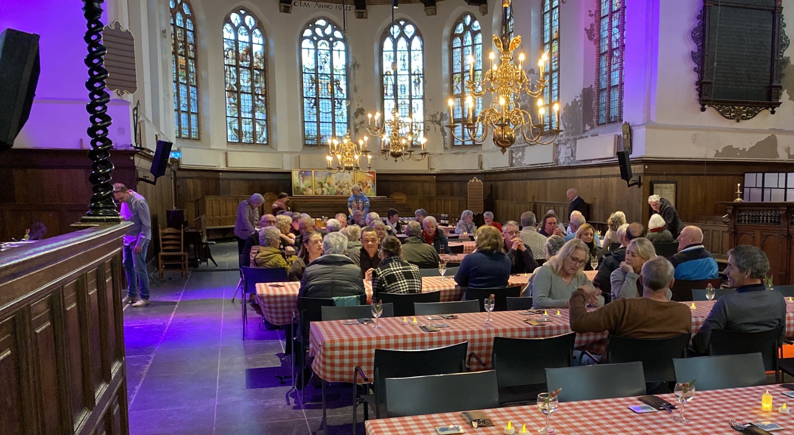 Groep mensen zittend aan tafels met geruite tafelkleden in een kerkzaal met glas-in-loodramen en kroonluchters.