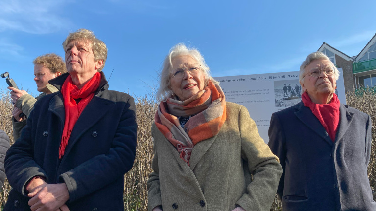 Drie mensen in dikke jassen en sjaals buiten op een zonnige dag. Op de achtergrond is een informatiebord te zien.