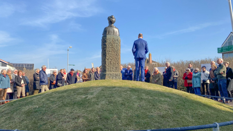 Een groep mensen verzameld rond een standbeeld op een grasheuvel onder een blauwe hemel.