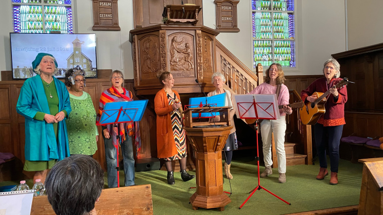 Zeven vrouwen zingen in een kerk, een van hen speelt gitaar; er is een houten preekstoel en een scherm met historische tekst.