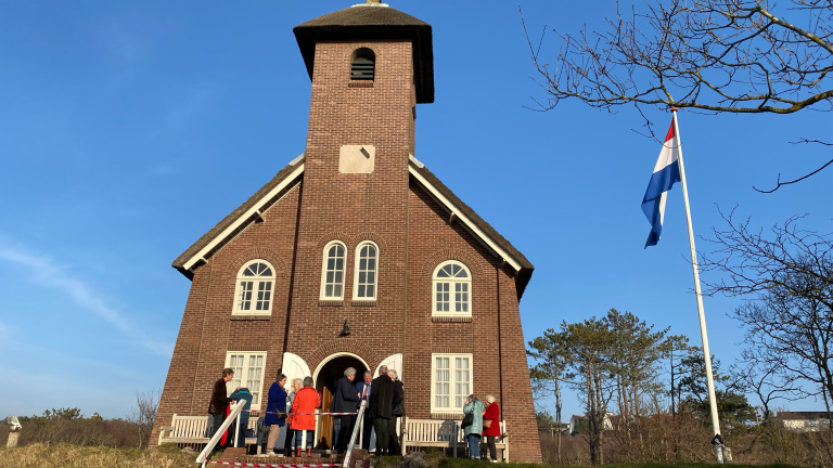 Bakstenen kerk met toren en mensen ervoor, naast een wapperende Nederlandse vlag.