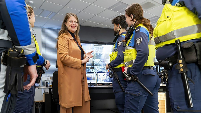 Een vrouw in een bruine jas spreekt met politieagenten in een controlekamer met beeldschermen op de achtergrond.