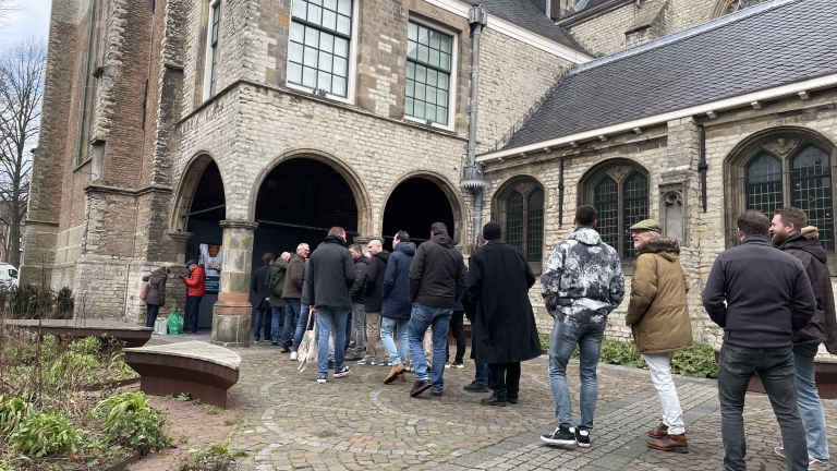 Mensen staan in de rij bij een stenen gebouw met bogen en glas-in-loodramen.