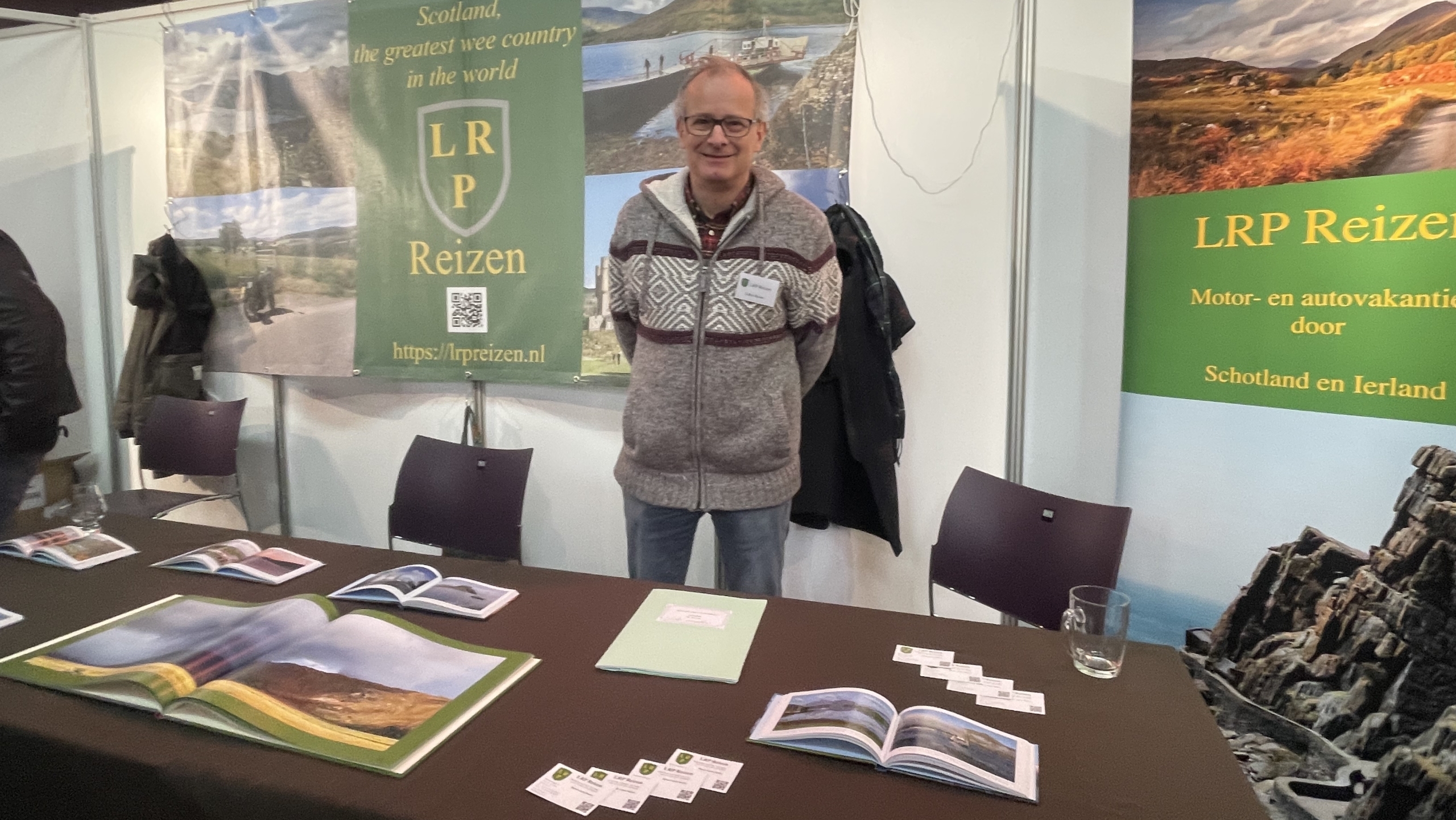 Een man staat voor een stand van LRP Reizen met affiches van Schotland en Ierland, een tafel met brochures en visitekaartjes.