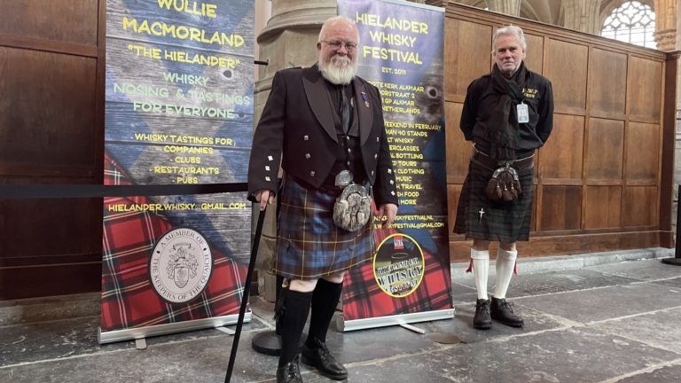 Twee mannen in Schotse kilts staan voor banners van het Hielander Whisky Festival.