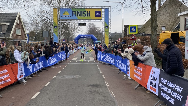 Een hardloper passeert de finishlijn van een wedstrijd, toegejuicht door een publiek langs de weg, met spandoeken van Mizuno en andere sponsoren.