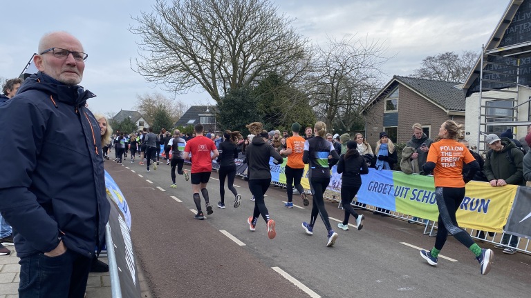 Een groep hardlopers tijdens een evenement met toeschouwers langs de zijlijn, en een man die hen bekijkt.