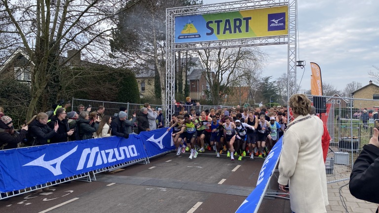 Begin van een hardloopwedstrijd met een grote groep renners die starten onder een boog met de tekst "Start", omringd door toeschouwers.