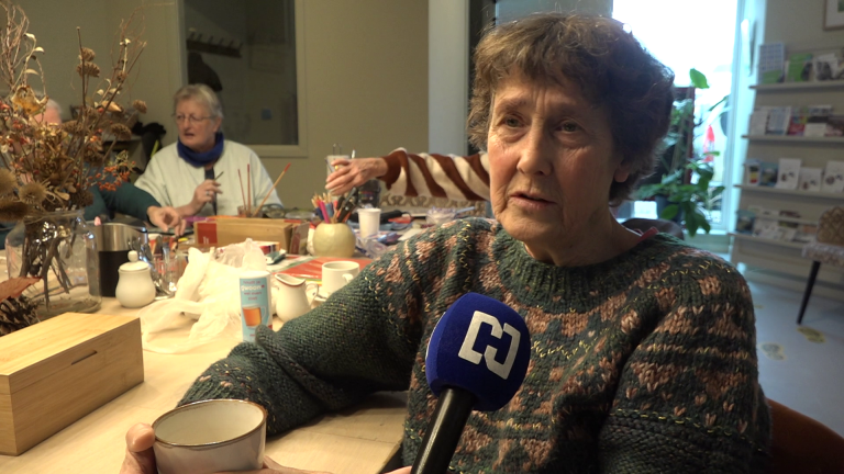 Oudere vrouw in een trui houdt een kopje vast en spreekt in een microfoon tijdens een creatieve activiteit aan tafel. Op de achtergrond zijn andere mensen aan het knutselen.