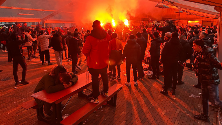 In een overdekte ruimte staan een grote groep mensen, sommige met fakkels, wat een rode gloed veroorzaakt. Op de voorgrond zit iemand met het hoofd op een tafel.