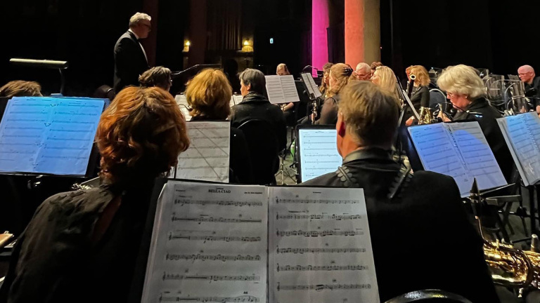 Orkestleden spelen muziek, gezien vanaf achter hun muzieklessenaars met bladmuziek, terwijl een dirigent voor hen staat.