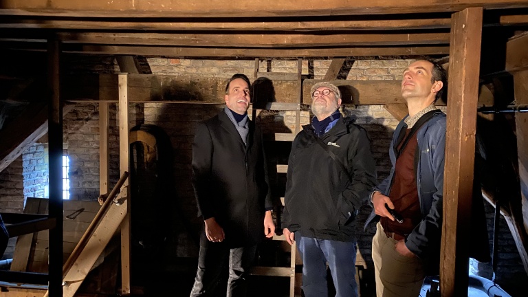 Drie mannen staan in een oude zolder met houten balken en bakstenen muren.