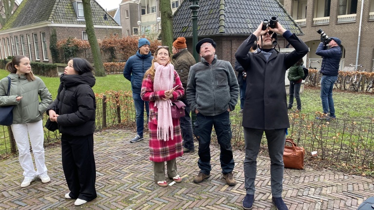 Groep mensen buiten, sommigen kijken omhoog met verrekijkers; beplanting en gebouw op de achtergrond.