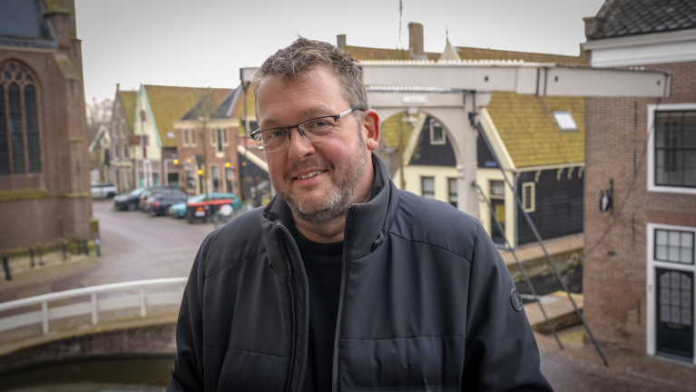 Een man met een bril en een donkere jas staat buiten met een historisch stadsbeeld op de achtergrond.