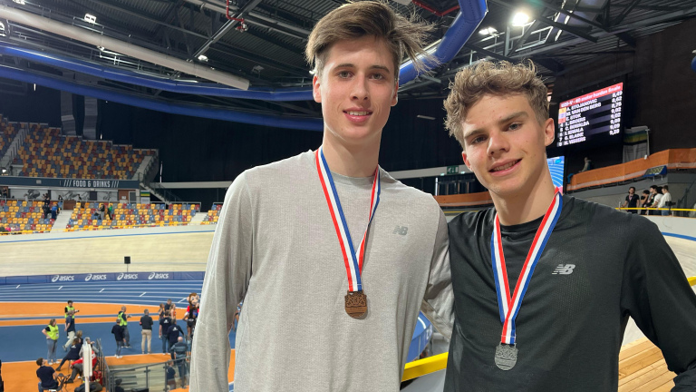 Twee jonge mannen met medailles poseren op een indoor atletiekbaan.