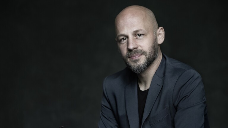 Kaalgeschoren man met een baard in een donker colbert, poserend voor een donkere achtergrond.