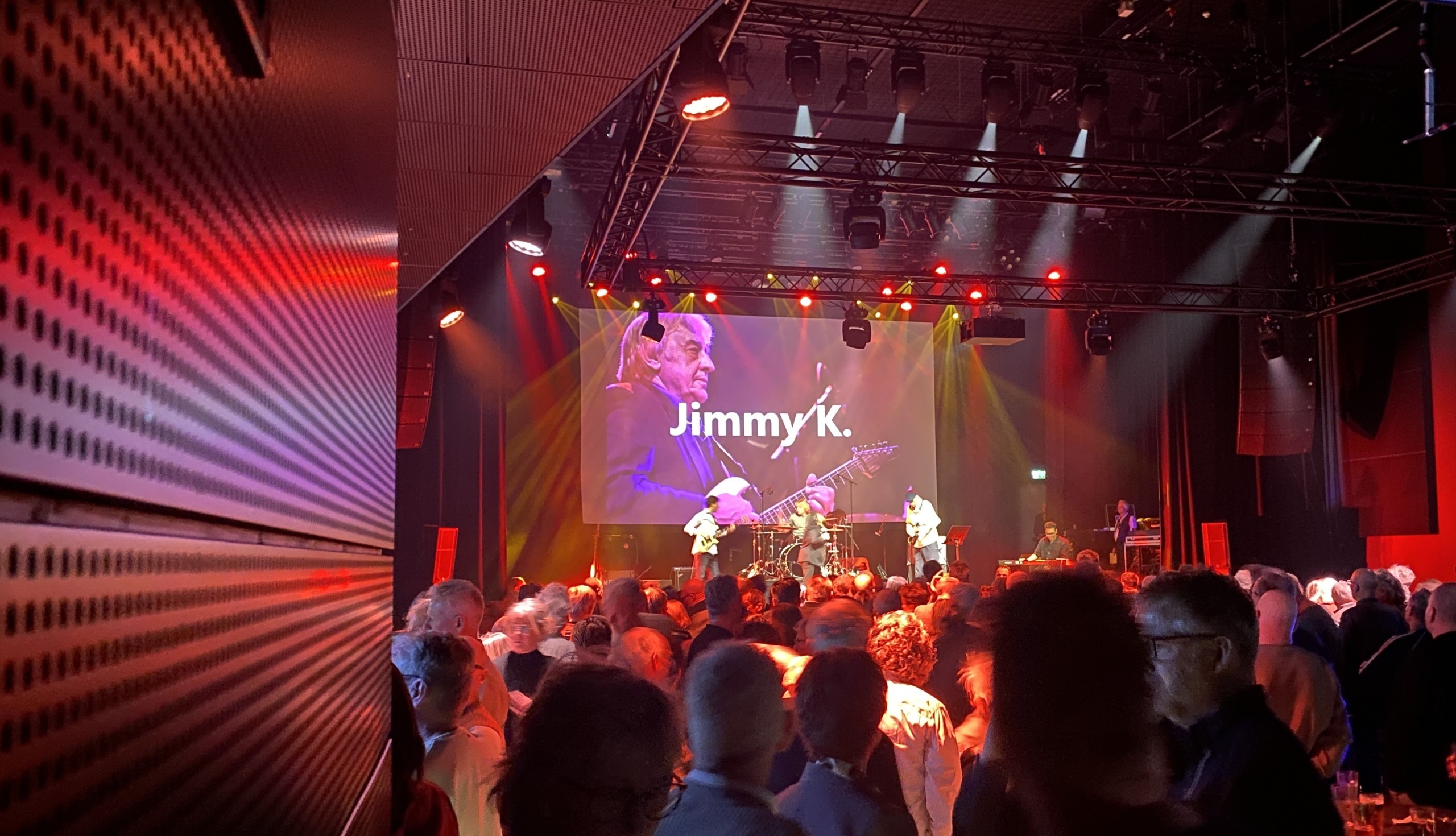 Mensen kijken naar een liveoptreden in een zaal met een groot scherm waarop "Jimmy K." wordt getoond.