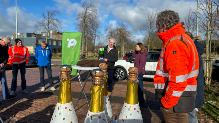 Groep mensen staat buiten in gesprek naast een tafel met versierde champagneflessen, met een groene vlag op de achtergrond.