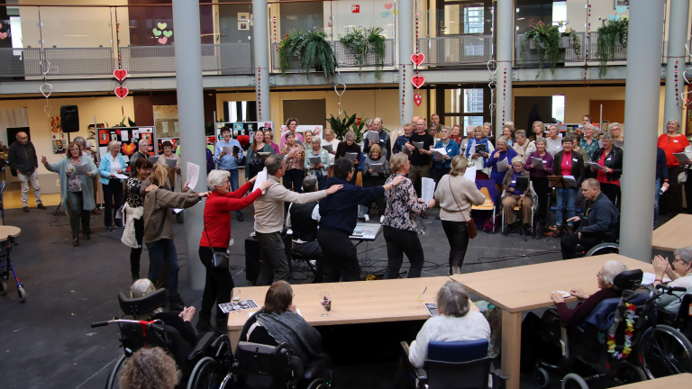 Groep mensen die zingen en muziek maken in een open binnenruimte, met publiek in rolstoelen en rondom tafels.