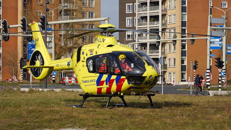 Een gele ambulancehelikopter op de grond met op de achtergrond een stadsomgeving en verkeerslichten.