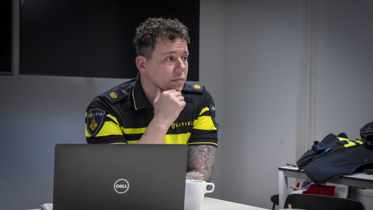 Man in politie-uniform, zittend aan een tafel, met een laptop en een kopje.