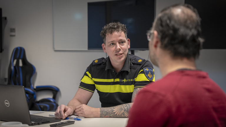 Een politieagent in uniform voert een gesprek aan tafel met een man in een rood shirt.