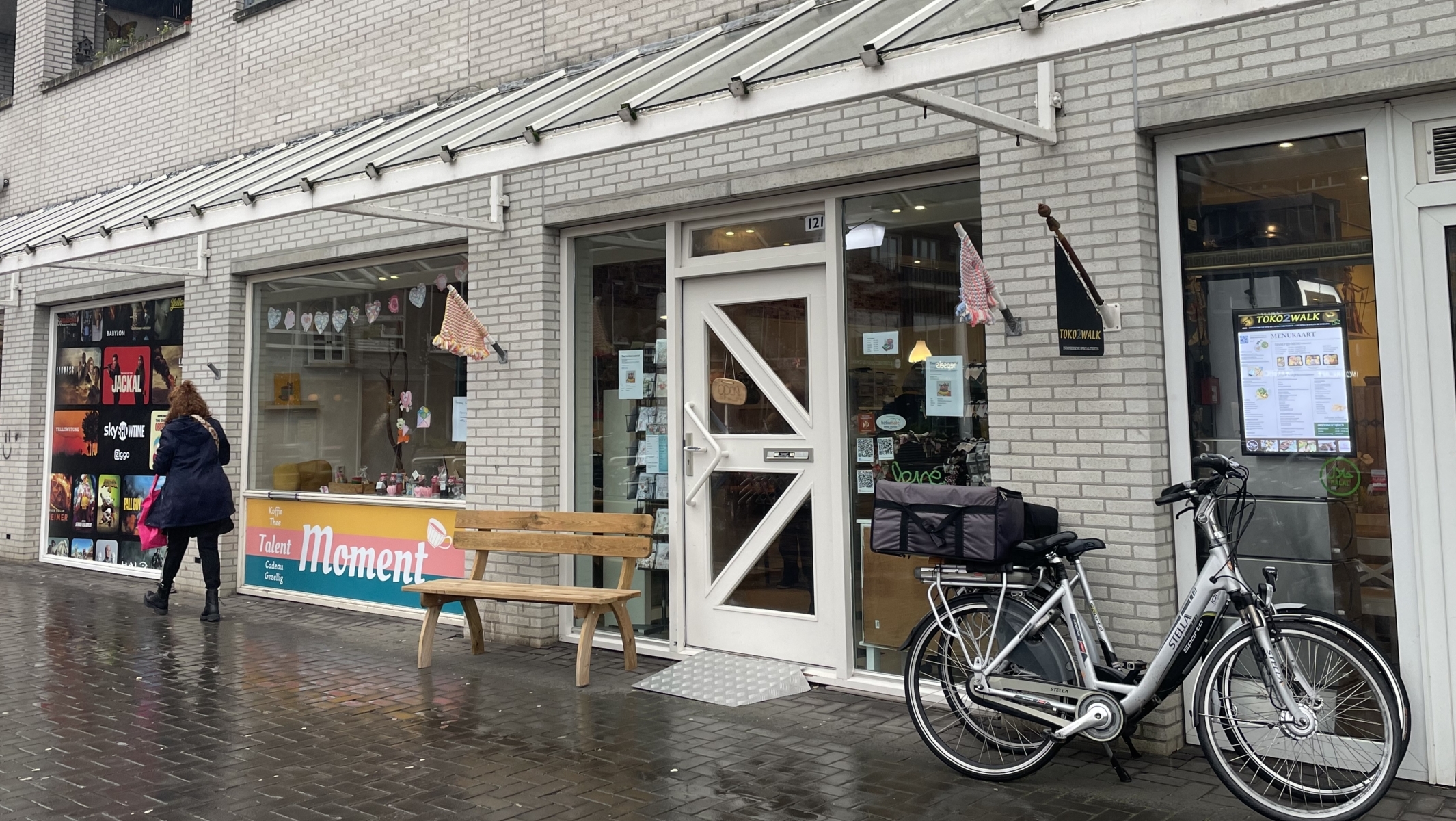 Een rij winkelpanden aan een straat met een fietser die langsloopt. Op de voorgrond staat een fiets geparkeerd voor de ingang van een winkel genaamd "TOKO 2." De straat is nat van de regen.