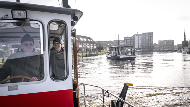 Twee mensen in de stuurhut van een boot op een rivier met een andere boot op de achtergrond en stedelijk landschap.