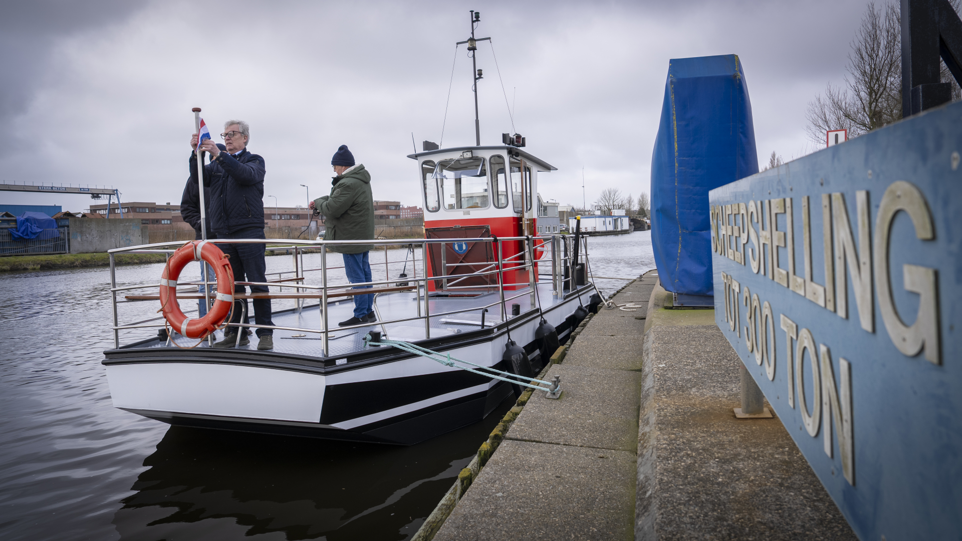 Twee mensen op een boot bij een dok, één hijst een vlag. Een bord met "scheepshelling tot 300 ton" is zichtbaar op de voorgrond.