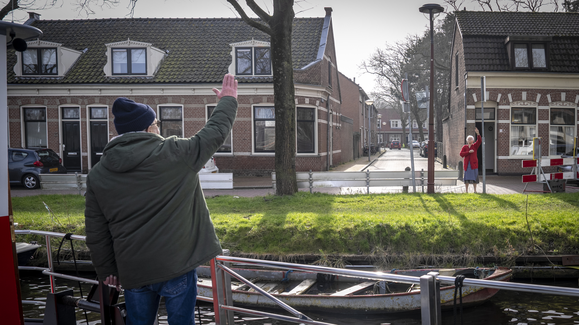 Man op een boot zwaait naar een vrouw die in een straat staat. Op de achtergrond zijn bakstenen huizen en een boom.