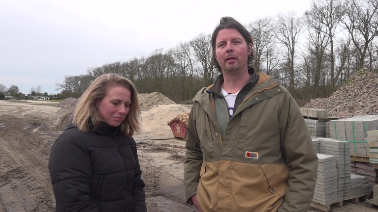 Twee mensen staan op een bouwterrein met zand en stapels stenen op de achtergrond.