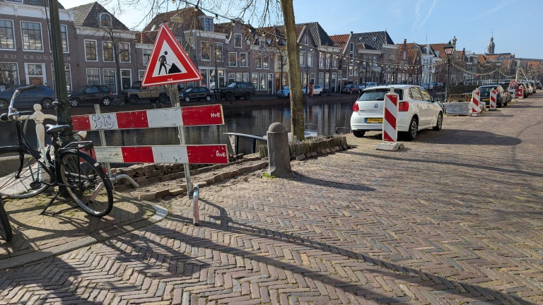 Straat met geparkeerde auto's bij een gracht, waarschuwingsbord en afzettingen bij stoepreparatie. Een fiets tegen een hek aan de linkerkant.