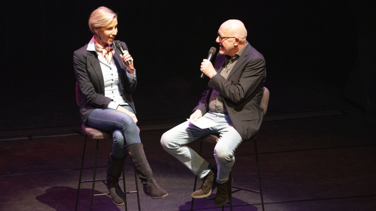 Twee mensen zitten op een podium en houden microfoons vast tijdens een gesprek.