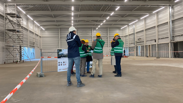 Een groep mensen met bouwhelmen en groene veiligheidshesjes staat in een grote, lege hal.