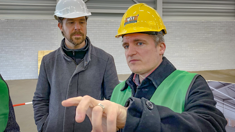 Twee mannen met veiligheidshelmen en groene hesjes in een industriële omgeving, wijzend naar iets buiten beeld.