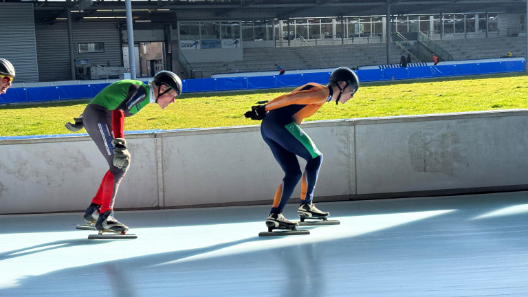 Twee schaatsers in felgekleurde pakken aan het schaatsen op een buitenbaan met gras op de achtergrond.