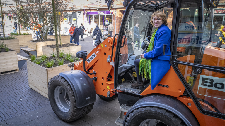 Vrouw zittend in een oranje graafmachine op straat, omringd door houten plantenbakken en voetgangers.