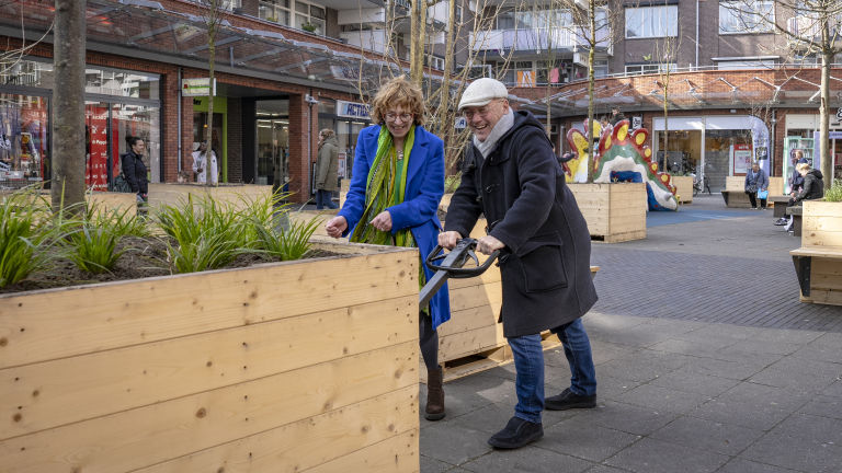 Twee mensen werken samen aan een grote houten plantenbak op een plein, omringd door andere plantenbakken en een kleurrijke sculptuur.