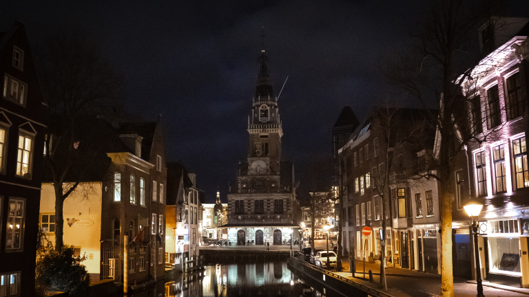 Nachtfoto van een grachtenpand in Nederland met een toren op de achtergrond, weerspiegeld in het water.