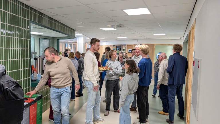 Groep mensen in een moderne lobby of kantine, sommigen drinken, terwijl anderen in gesprek zijn; een man biedt hapjes aan op een schaal.