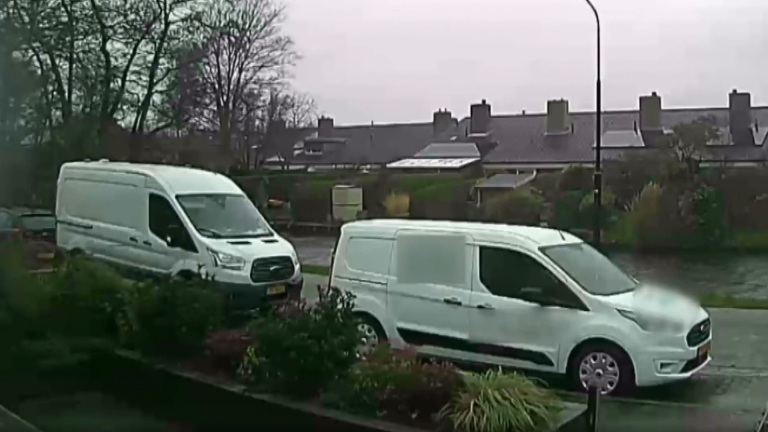 Twee witte bestelwagens geparkeerd langs een straat met huizen op de achtergrond en kale bomen.