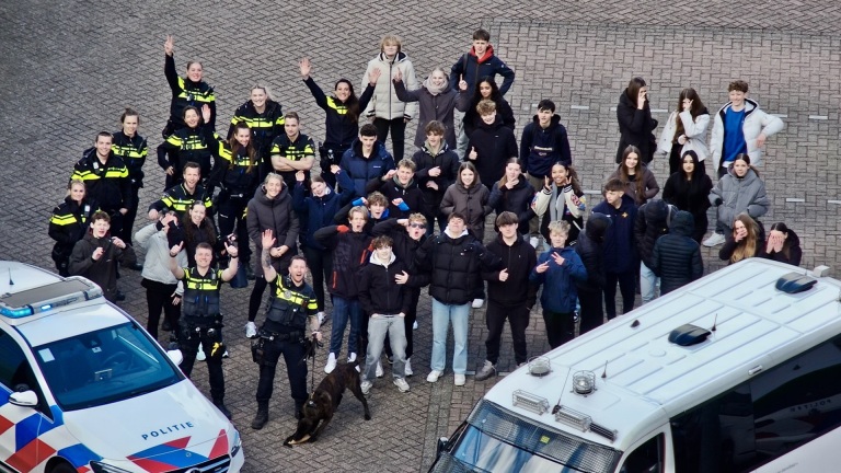 Groep mensen, waaronder politieagenten en jongeren, staan buiten naast politievoertuigen en zwaaien naar de camera.