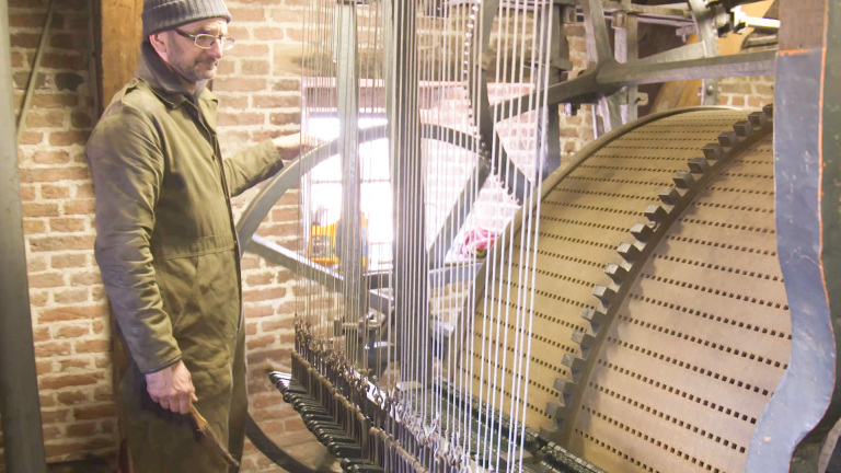 Een man met een muts en bril staat naast een groot mechanisme met verticale draden en een grote, perforated draaiende trommel in een ruimte met bakstenen muren.