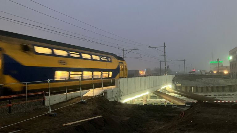 Trein rijdt over een verhoogde spoorweg met bouwplaats ernaast in de avondschemering, omgeven door nevelige lucht.