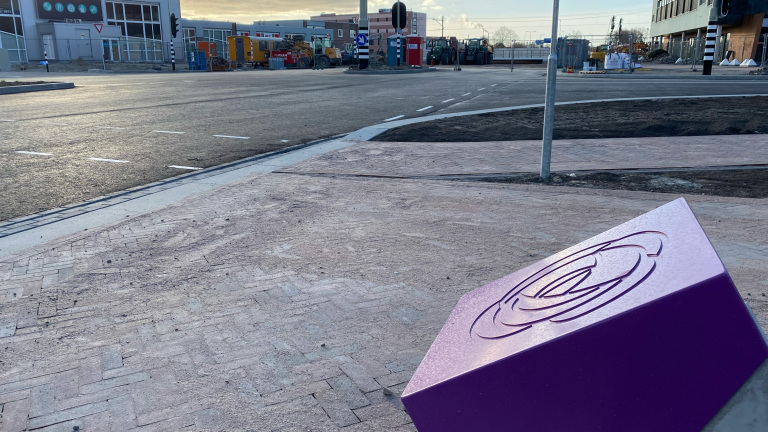Straatbeeld met een paars kunstobject op de voorgrond en een nieuwe weg in aanbouw op de achtergrond, met bouwmachines en gebouwen zichtbaar in de verte.