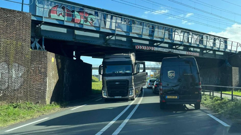 Vrachtwagen die vastzit onder een lage spoorbrug met een UPS-bestelbusje ervoor op de weg; de brug is voorzien van graffiti.