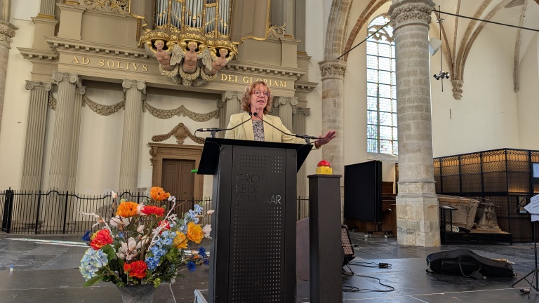 Een persoon staat achter een spreekgestoelte in de Grote Kerk van Alkmaar, omgeven door architectonische details en een boeket kleurrijke bloemen op de voorgrond.