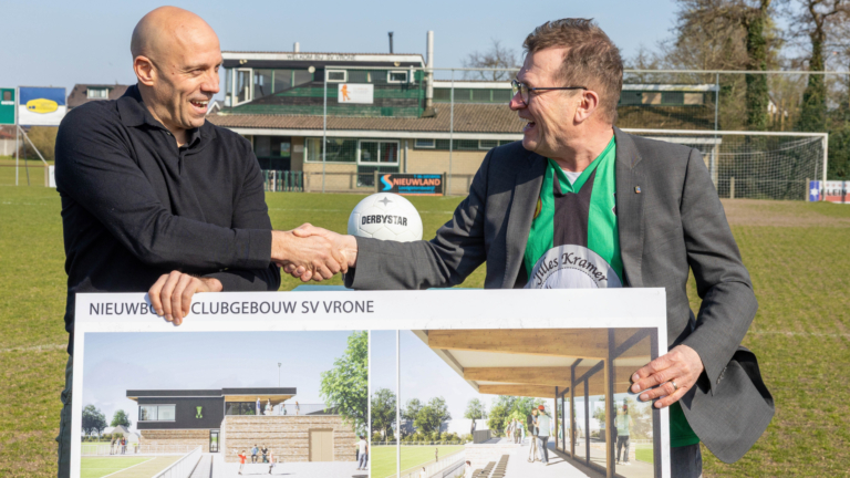Twee mannen schudden elkaar de hand op een voetbalveld, terwijl ze een ontwerpplan voor het nieuwe clubgebouw van SV Vrone vasthouden.